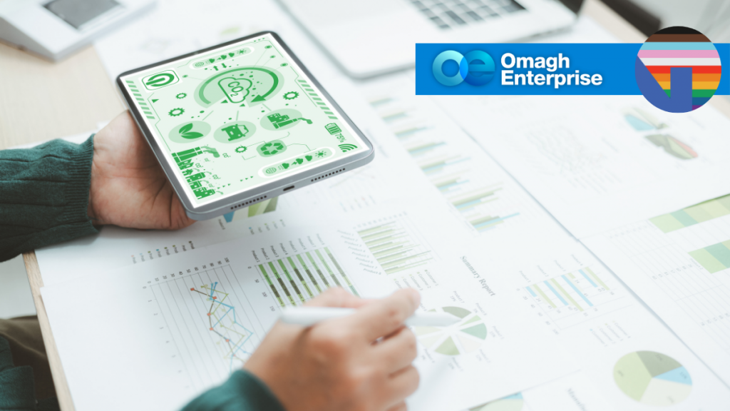 A person holds a tablet displaying green analytics graphics while reviewing printed charts and graphs on a desk. The Omagh Enterprise logo and a rainbow-striped circular icon appear in the top right corner.