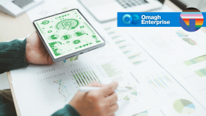 A person holds a tablet displaying green analytics graphics while reviewing printed charts and graphs on a desk. The Omagh Enterprise logo and a rainbow-striped circular icon appear in the top right corner.