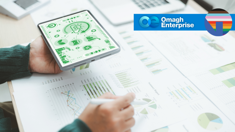 A person holds a tablet displaying green analytics graphics while reviewing printed charts and graphs on a desk. The Omagh Enterprise logo and a rainbow-striped circular icon appear in the top right corner.