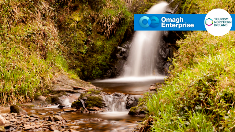 A small waterfall flows over rocks surrounded by green grass and moss; logos for Omagh Enterprise and Tourism Northern Ireland appear in the top right corner.