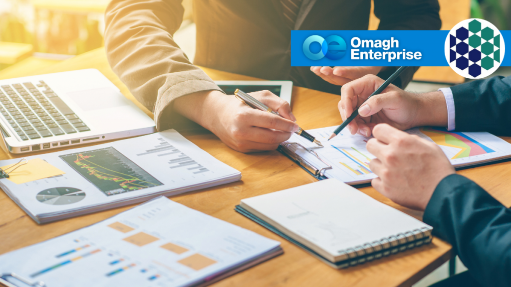 Two people review financial charts and graphs at a desk with a laptop. The Omagh Enterprise logo appears in the top right corner.