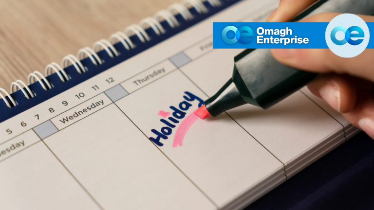 A hand uses a pink highlighter to mark the word Holiday written in blue on a calendar. The logo Omagh Enterprise appears in the top right corner.