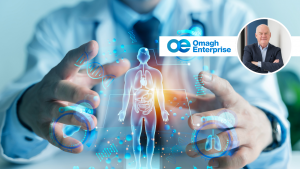 A doctor in a white coat uses a digital interface displaying a human body and medical icons. The Omagh Enterprise logo and a small photo of a man in a suit appear in the top right corner.