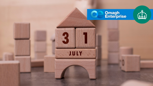 Wooden blocks arranged to form a house display the date 31 July. In the top right corner, there is a blue banner with the Omagh Enterprise logo and a crown icon on a green circle.