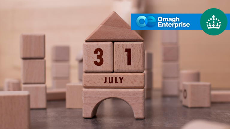 Wooden blocks arranged to form a house display the date 31 July. In the top right corner, there is a blue banner with the Omagh Enterprise logo and a crown icon on a green circle.