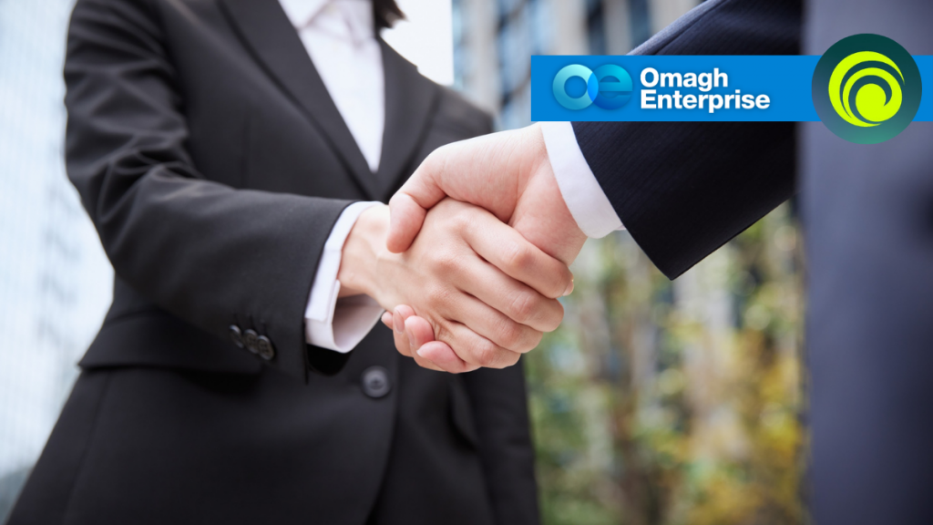 Two people in business attire shaking hands outdoors, with the Omagh Enterprise logo and branding visible in the upper right corner.