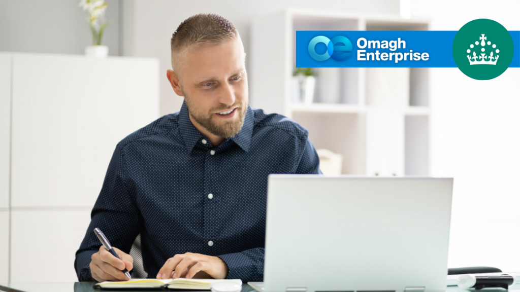 Man sitting at a desk, writing in a notebook while looking at a laptop; Omagh Enterprise and a crown logo are visible in the top right corner.