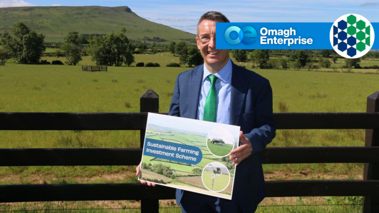 DAERA Minister Andrew Muir in a suit stands outside holding a "Sustainable Farming Investment Scheme" sign in front of a field, with the Omagh Enterprise logo in the top right corner.