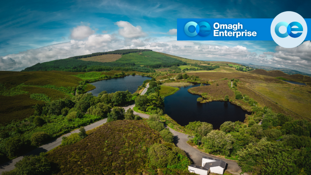 Aerial view of a hilly landscape with lakes, greenery, and roads under a blue sky; Omagh Enterprise logo in the top right corner.