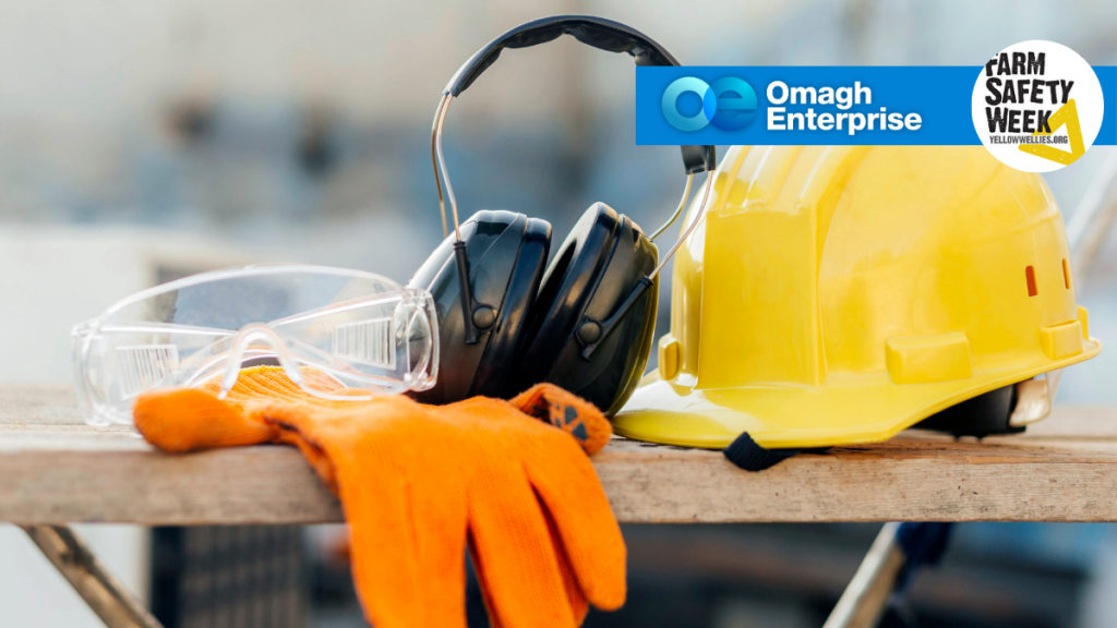 Safety gear including a hard hat, gloves, goggles, and ear protection on a workbench. Omagh Enterprise and Farm Safety Week logos are visible in the background.