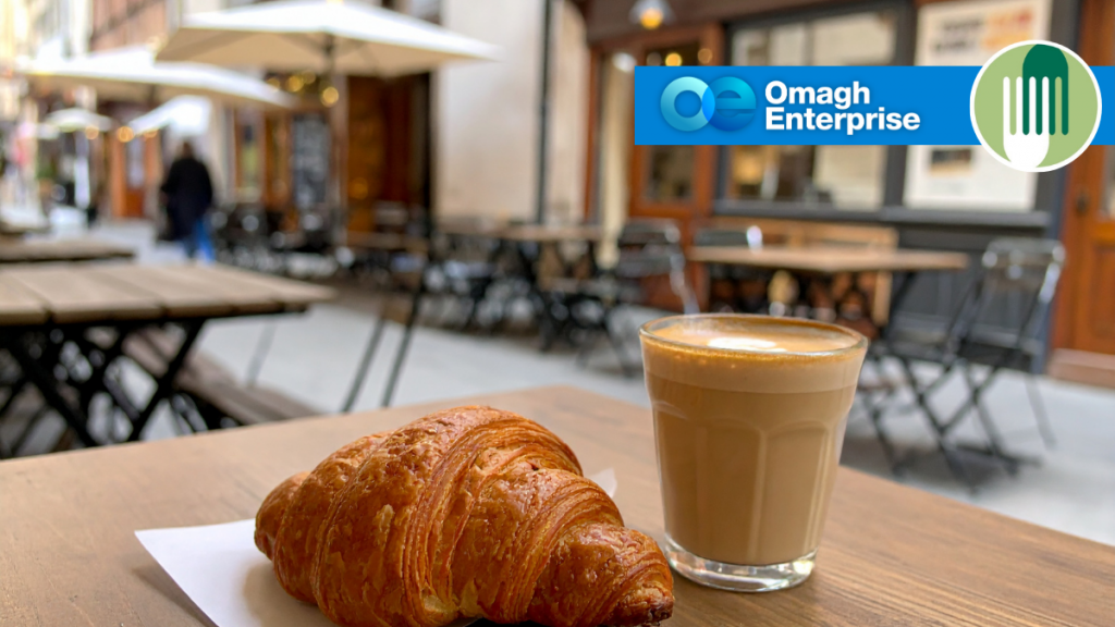 A croissant and a glass of coffee sit on a table at an outdoor café. The Omagh Enterprise logo and another green logo appear in the top right corner.