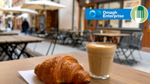 A croissant and a glass of coffee sit on a table at an outdoor café. The Omagh Enterprise logo and another green logo appear in the top right corner.