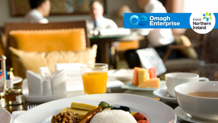 A breakfast with rice, vegetables, juice, and fruit sits on a table; two people sit blurred in the background. Logos for Omagh Enterprise and Invest Northern Ireland are in the top right corner.