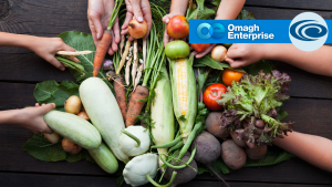 Several hands reach toward a variety of fresh vegetables, including carrots, zucchini, tomatoes, and lettuce, arranged on a wooden table. "Omagh Enterprise" logo appears in the top right.