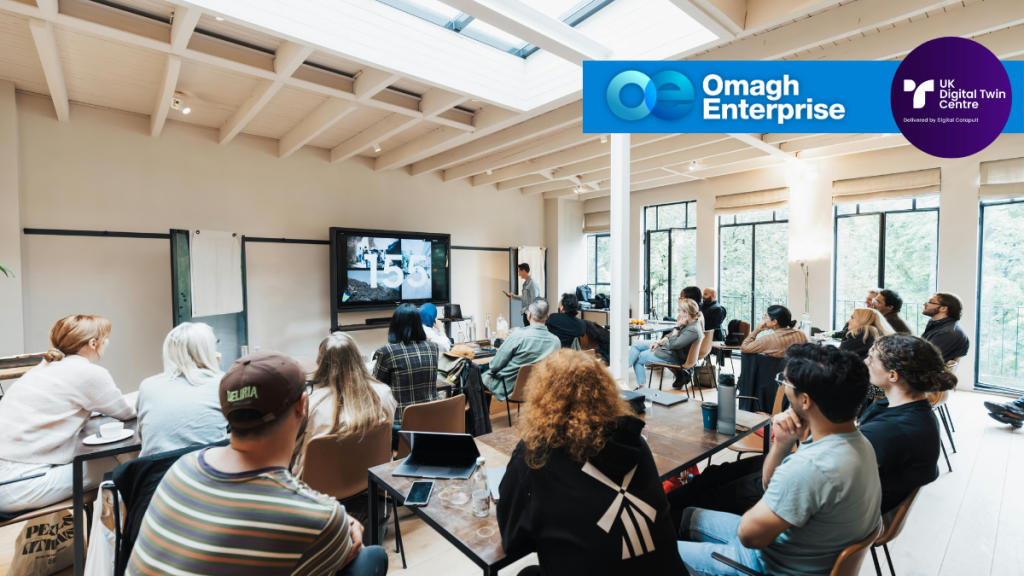 A group of people in a modern, well-lit meeting room watch a speaker give a presentation; logos for Omagh Enterprise and UK Digital Twin Centre are displayed.