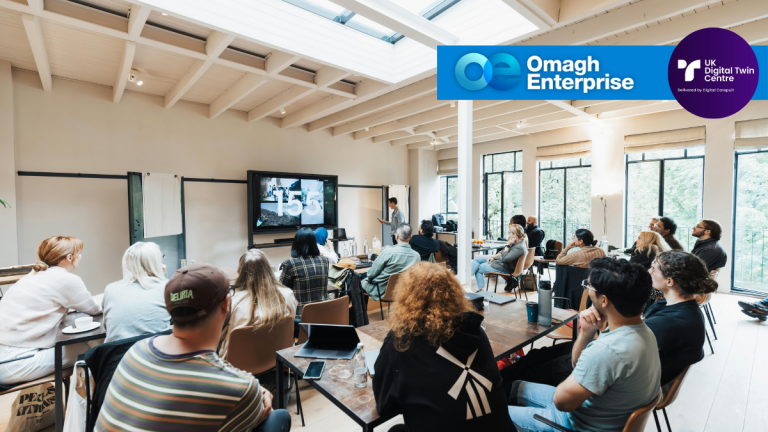 A group of people in a modern, well-lit meeting room watch a speaker give a presentation; logos for Omagh Enterprise and UK Digital Twin Centre are displayed.