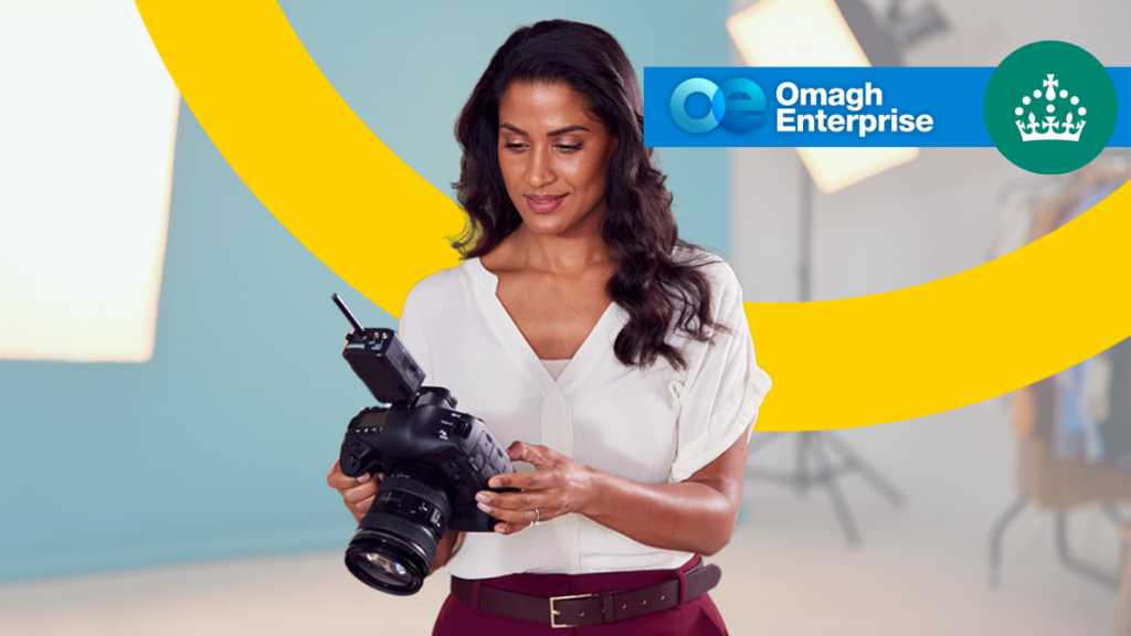 A woman in a white blouse looks down at a professional camera. Behind her are bright studio lights, a yellow swoosh, and Omagh Enterprise and government logos.