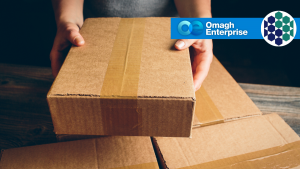 Person holding a cardboard box over two other boxes, with the Omagh Enterprise logo in the top right corner.