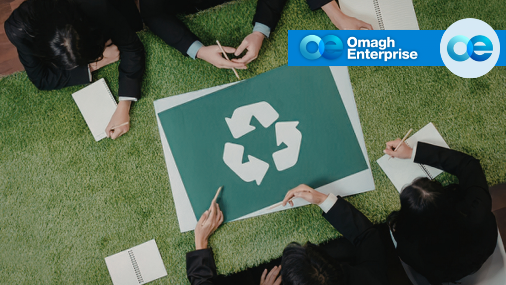 Four people sit around a table with a recycling symbol on it, taking notes. The Omagh Enterprise logo is displayed in the upper right corner.