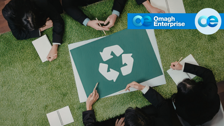 Four people sit around a table with a recycling symbol on it, taking notes. The Omagh Enterprise logo is displayed in the upper right corner.