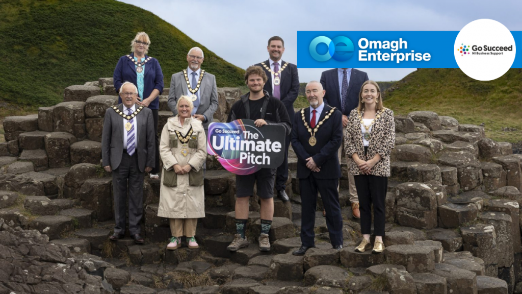 A group of people stand on Giant’s Causeway rocks, some wearing ceremonial chains, holding a sign reading “Go Succeed The Ultimate Pitch.” Omagh Enterprise and Go Succeed logos are in the top right.