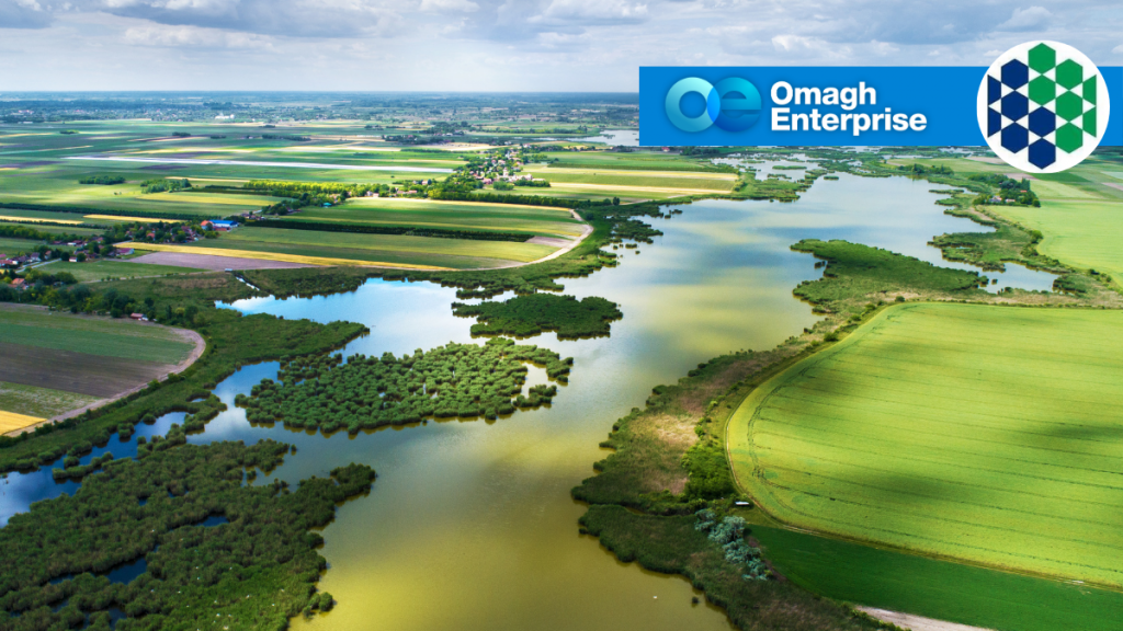 Aerial view of green fields and a winding river under a cloudy sky, with the Omagh Enterprise logo superimposed in the top right corner.