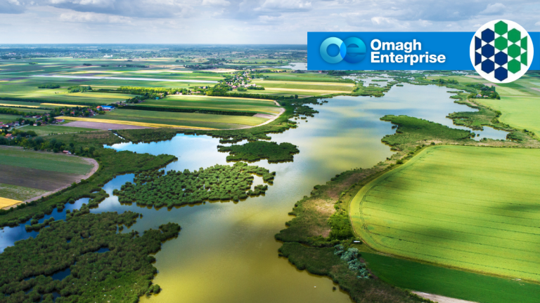 Aerial view of green fields and a winding river under a cloudy sky, with the Omagh Enterprise logo superimposed in the top right corner.