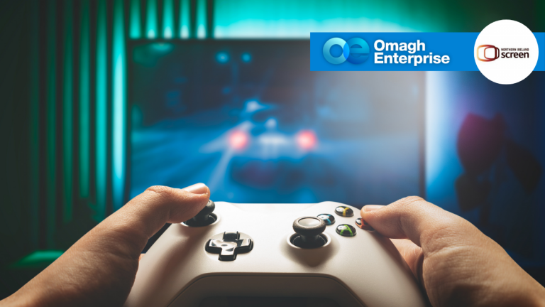 Close-up of hands holding a game controller in front of a blurred racing video game on a screen, with Omagh Enterprise and National Lottery Open Fund logos in the corner.