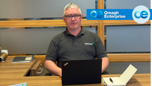 Sean Donnelly sits at a desk with a laptop, notepad, and pen. The Omagh Enterprise logo is visible in the background.