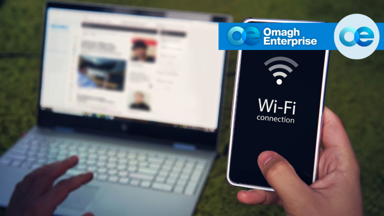 A person uses a smartphone showing “Wi-Fi connection” in front of a blurred laptop screen. “Omagh Enterprise” logo is displayed in the corner.