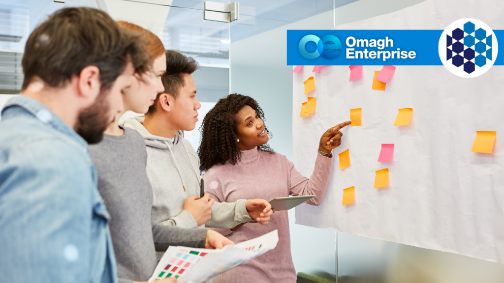 Four people stand by a whiteboard covered in sticky notes, discussing ideas. One woman points to the board. Omagh Enterprise logo is visible in the corner.