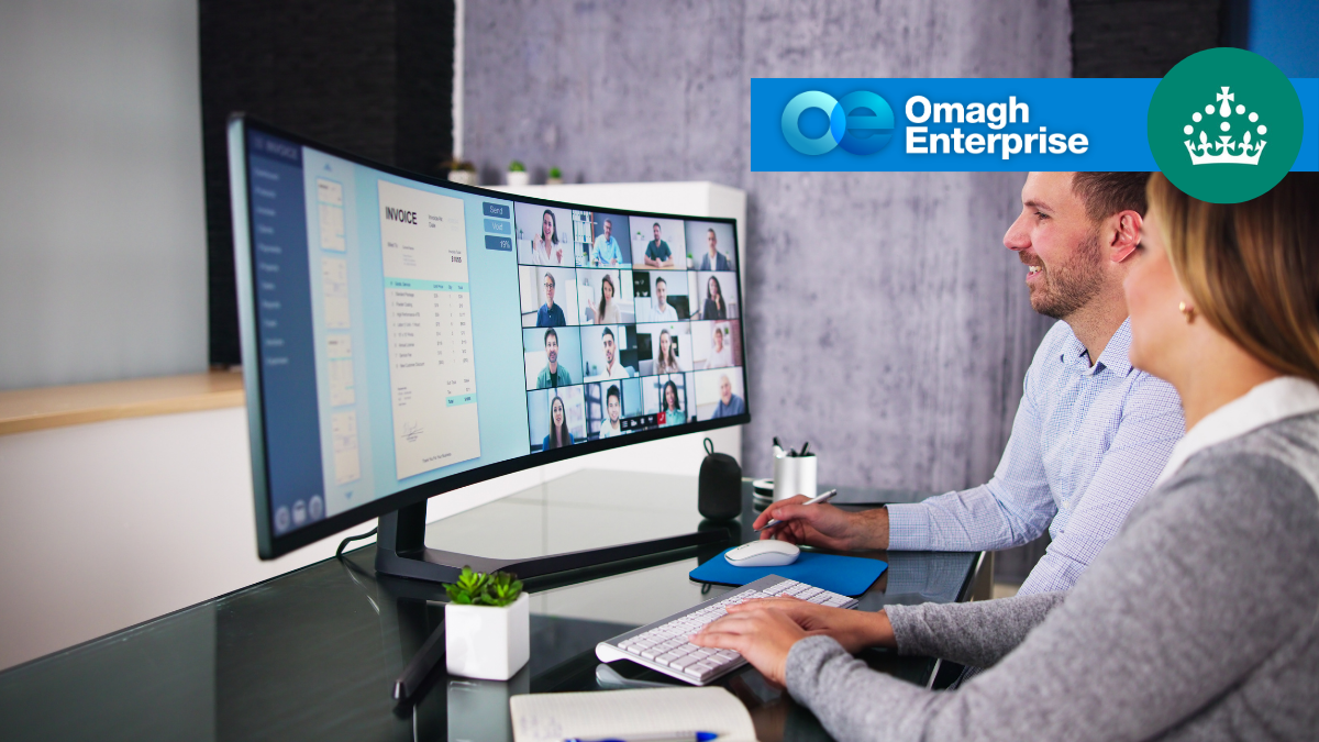 Two people sit at a desk and participate in a group video conference on a wide curved monitor. The Omagh Enterprise and Crown logos are displayed in the upper right corner.