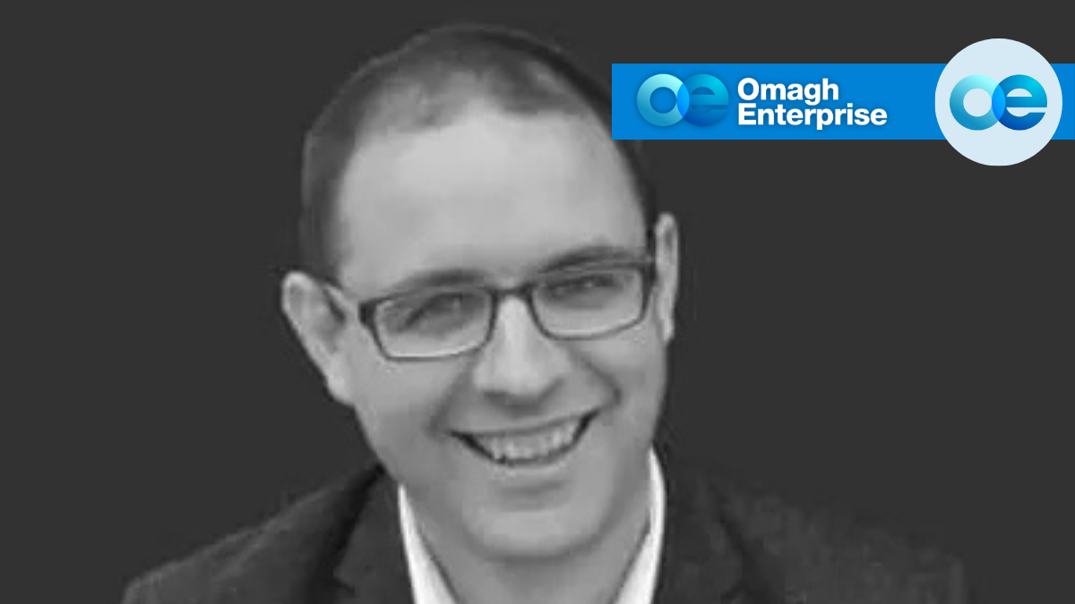Smiling man wearing glasses and a suit jacket; Omagh Enterprise logo appears in the top right corner on a dark background.