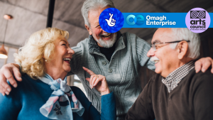 Three older adults sit together indoors, smiling and laughing. Logos for Omagh Enterprise, the Arts Council of Northern Ireland, and the National Lottery are displayed at the top of the image.
