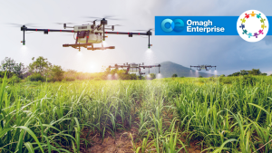 Drones spray crops in a green field with hills in the background, under a cloudy sky. Omagh Enterprise and a circular logo appear in the top right corner.