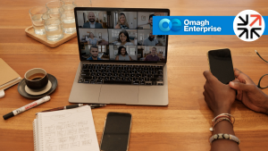 A laptop on a wooden table displays a group video call, with a person using a smartphone beside notebooks, pens, drinks, and Omagh Enterprise and business logos.