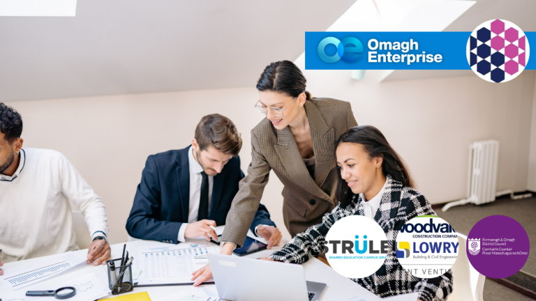 Four people work at a table with laptops and papers; a woman stands and points at a screen. Logos for Omagh Enterprise and other organizations are displayed in the foreground.