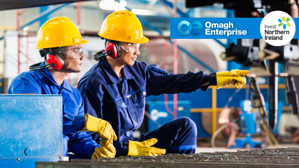 Two workers in blue uniforms and yellow helmets operate machinery in a factory. The Omagh Enterprise and Invest Northern Ireland logos appear in the upper right corner.