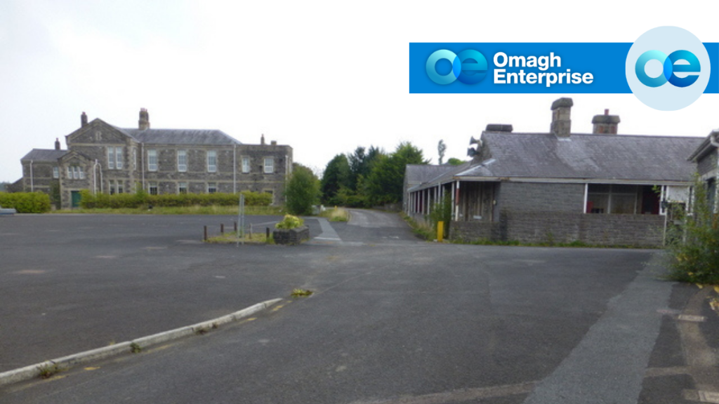 Two stone buildings with chimneys stand in a mostly empty paved area under a cloudy sky. An “Omagh Enterprise” logo appears in the top right corner.