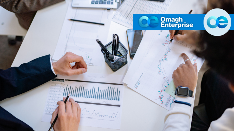 Three people review financial charts and graphs at a table. The Omagh Enterprise logo appears in the top right corner of the image.