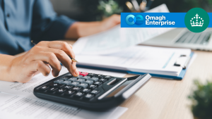 A person using a calculator at a desk with paperwork and a laptop, overlaid with the Omagh Enterprise logo.