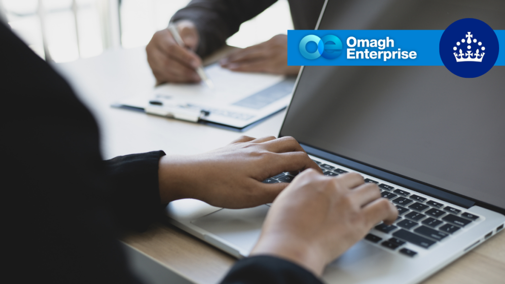 A person typing on a laptop with another person reviewing documents in the background, overlaid with the Omagh Enterprise logo.