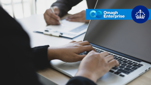 A person typing on a laptop with another person reviewing documents in the background, overlaid with the Omagh Enterprise logo.