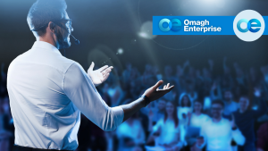 A speaker wearing a headset gestures to a blurred audience, with the Omagh Enterprise logo in the upper right.