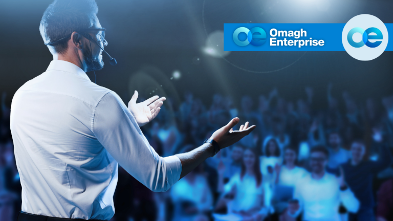 A speaker wearing a headset gestures to a blurred audience, with the Omagh Enterprise logo in the upper right.