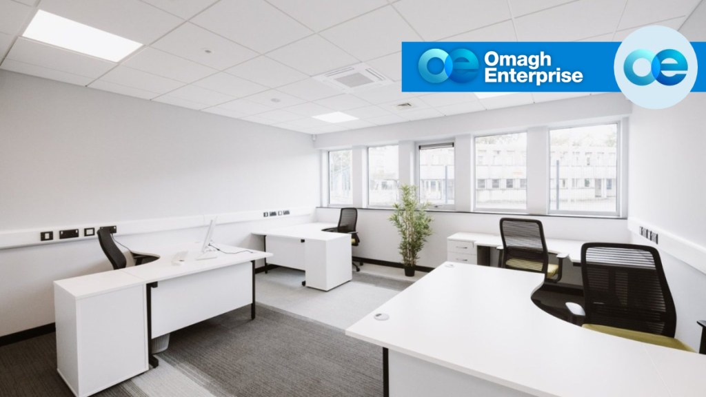 Empty office space with white desks, black chairs, and windows, featuring an Omagh Enterprise logo in the corner.