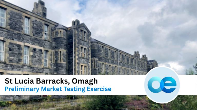 A large, old stone building labeled St Lucia Barracks, Omagh, with cloudy skies above and overgrown vegetation in the foreground.