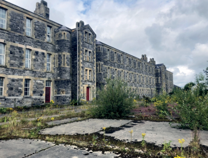 The former St Lucia Barracks buildings in Omagh, with grass and overgrowth in the foreground."